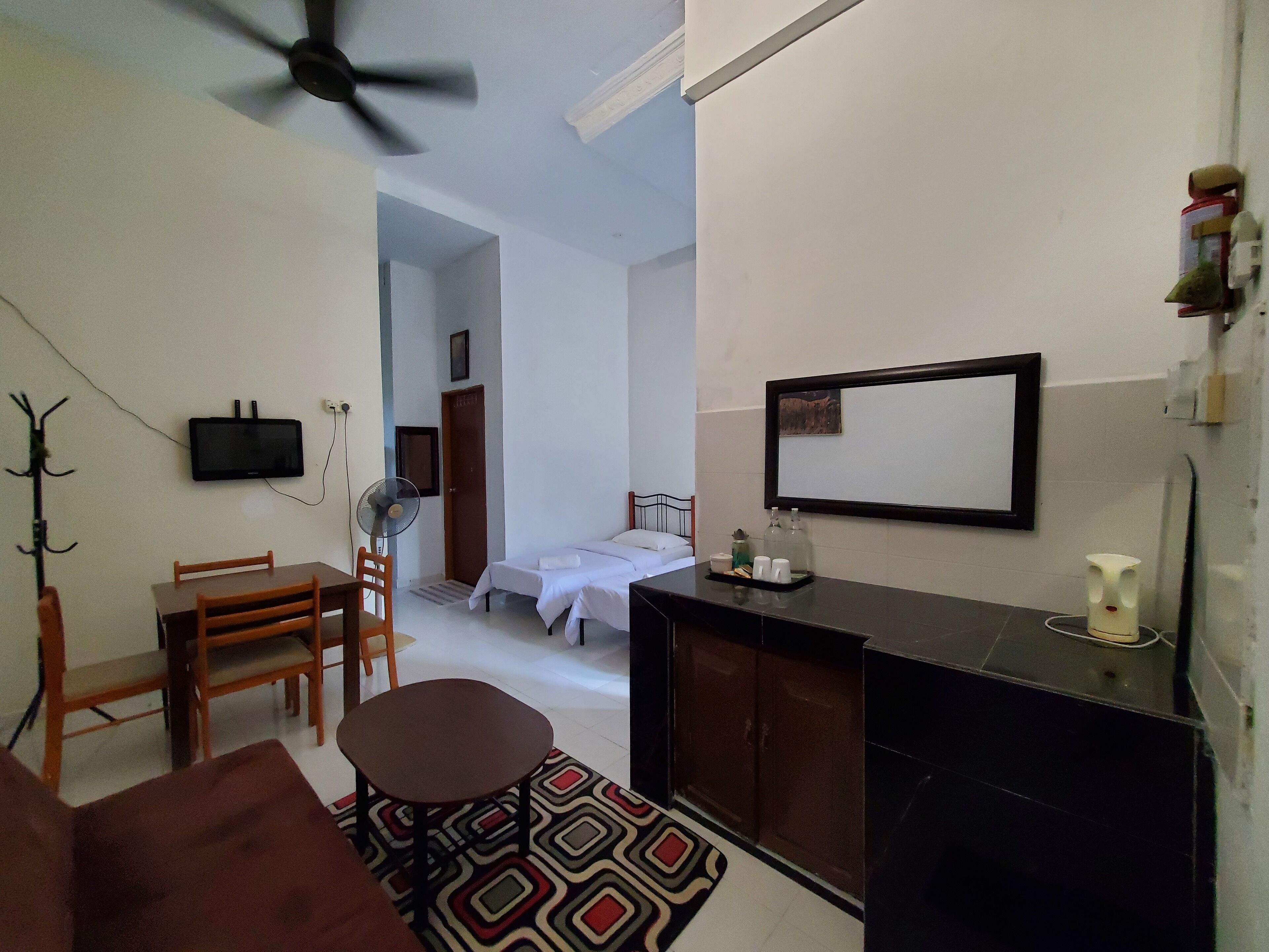 Ein kleines Hotelzimmer mit Bett, Schreibtisch, Stuhl, Nachttisch, einer Sitzecke, Deckenventilator, Fernseher, Spiegel und einem gemusterten Teppich.