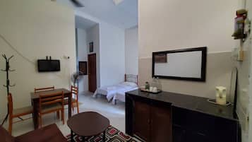 Ein kleines Hotelzimmer mit Bett, Schreibtisch, Stuhl, Nachttisch, einer Sitzecke, Deckenventilator, Fernseher, Spiegel und einem gemusterten Teppich.