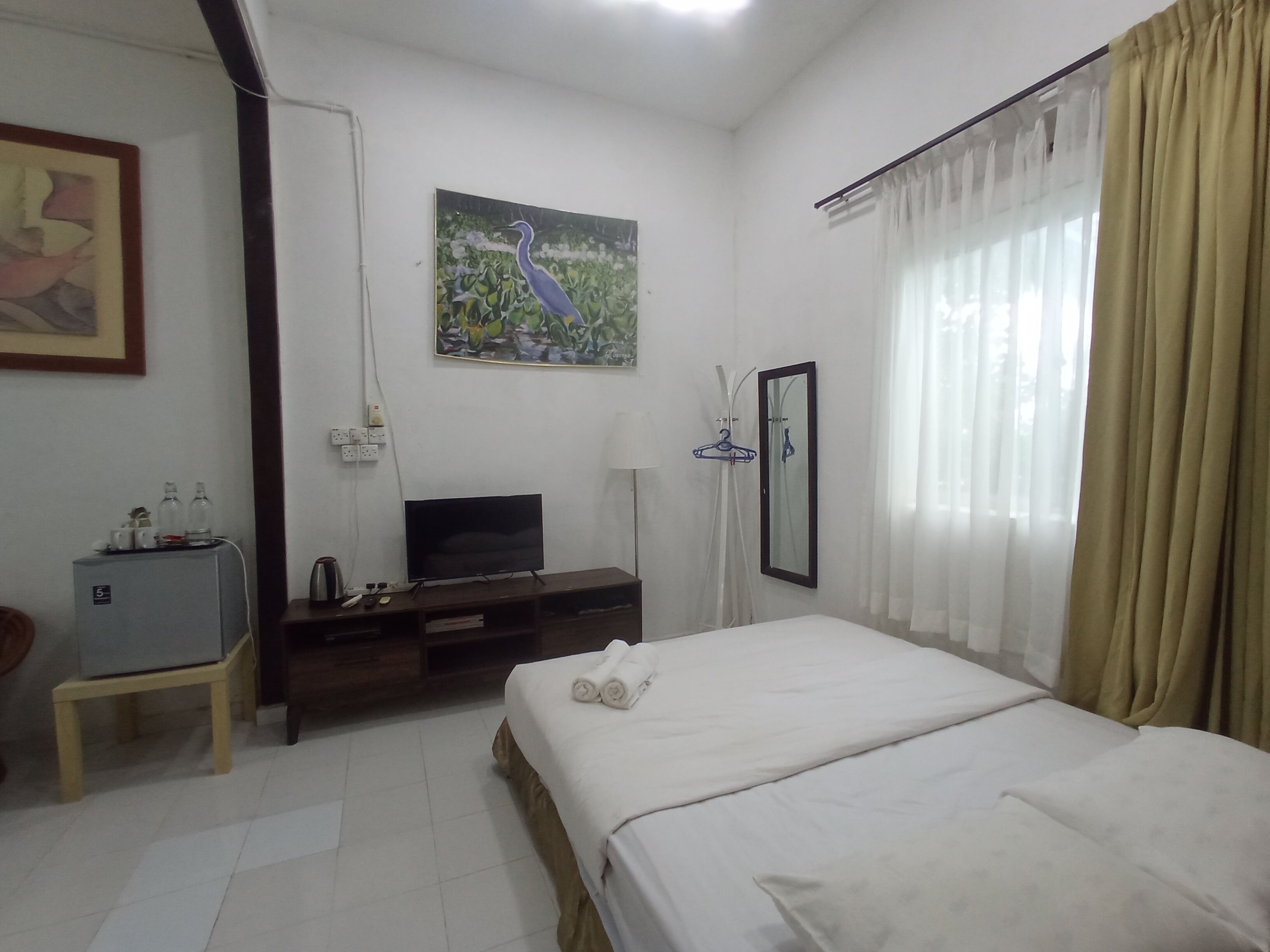 A hotel room with a bed, a TV, a small cabinet, a painting, and a window with curtains.