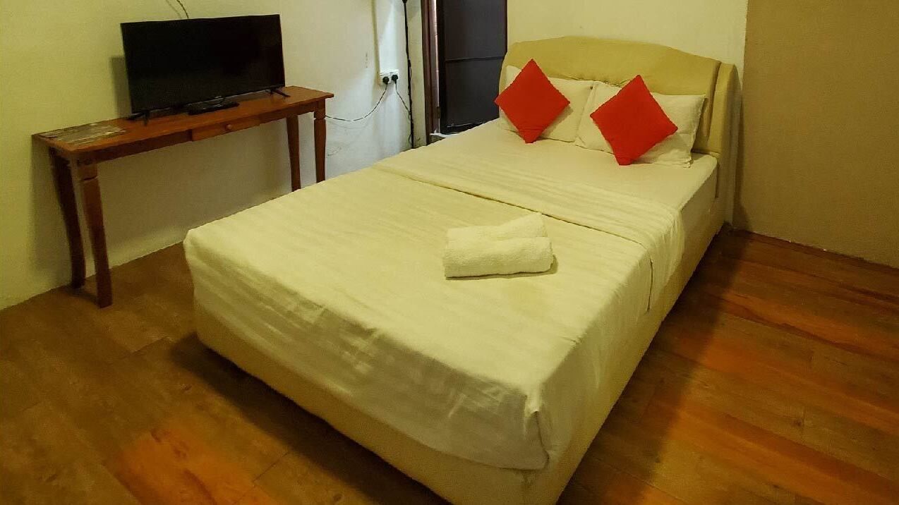 A neatly made bed with a white bedspread and two red pillows, a wooden nightstand with a television, and a plain wall.