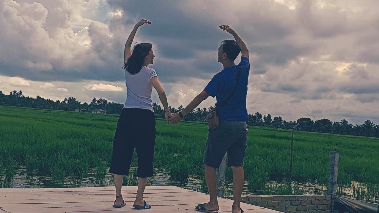 Dos personas tomadas de la mano sobre un muelle de madera, con un campo verde y un cielo nublado al fondo.