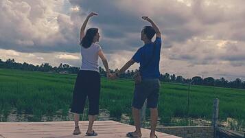Dos personas tomadas de la mano sobre un muelle de madera, con un campo verde y un cielo nublado al fondo.