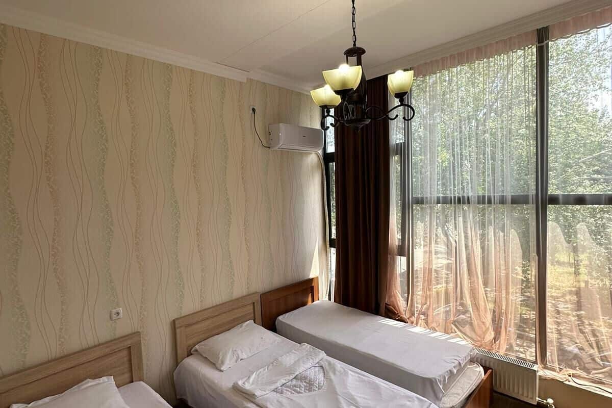 A hotel room with two single beds, a chandelier, a large window with curtains, and a wall-mounted air conditioner.
