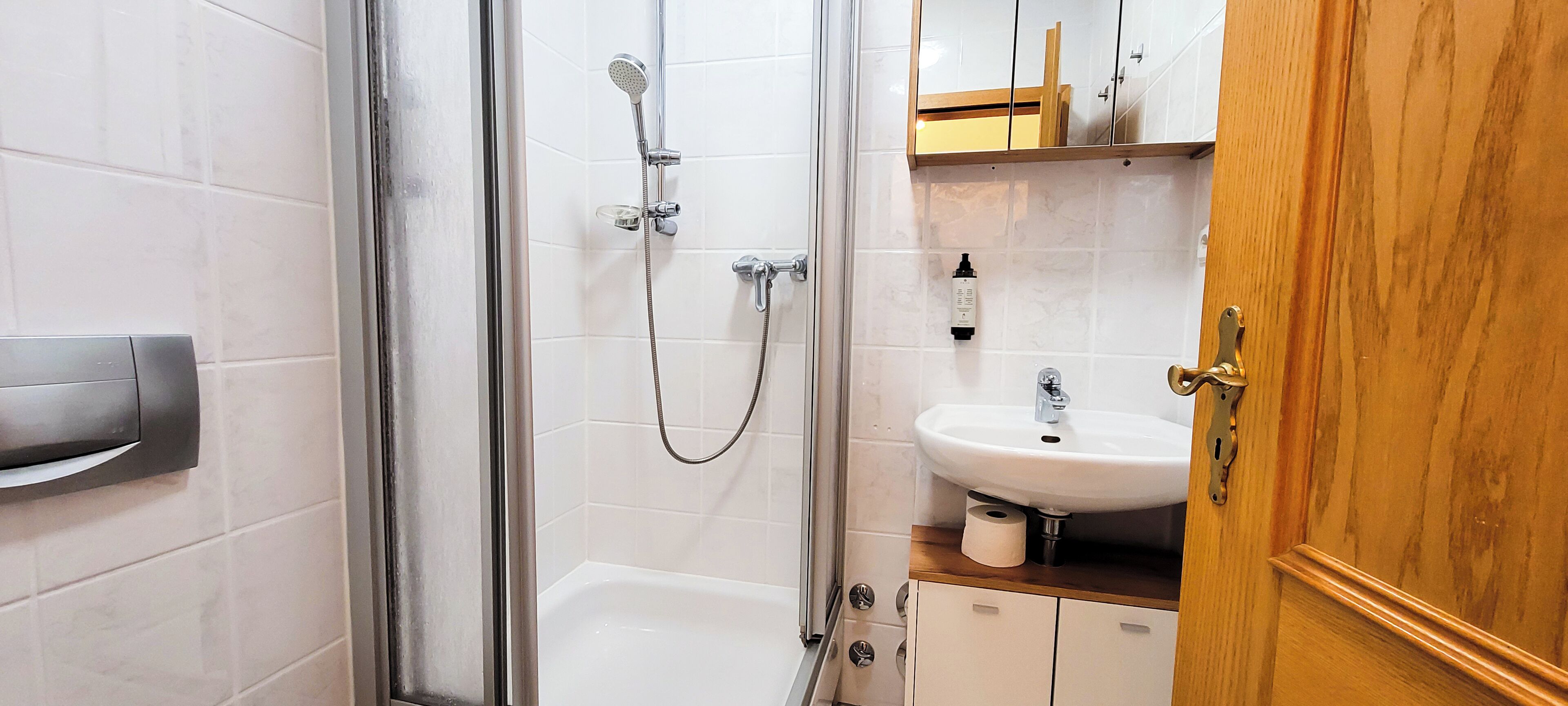 A bathroom with a glass shower enclosure, a white sink, and wooden cabinets.