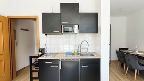 Een kleine keuken met een magnetron, spoelbak en zwarte kastjes. Er is een eethoek met een tafel voor twee personen.