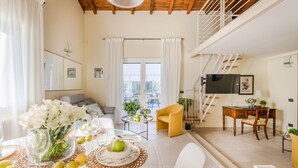 En rummelig opholdsstue med højt til loftet, et spisebord dækket med frugt og glas, en sofa, og en trappe op til et øvre plan.
