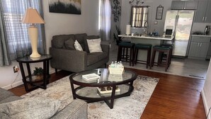 Ein Wohnzimmer mit grauem Sofa, einem runden Couchtisch und einer Küchenzeile inklusive Kühlschrank und Barhocker.