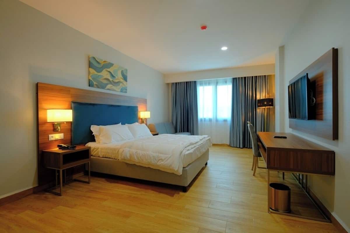 Una habitación de hotel con cama, mesitas de noche, escritorio, silla, televisión y ventana con cortinas.