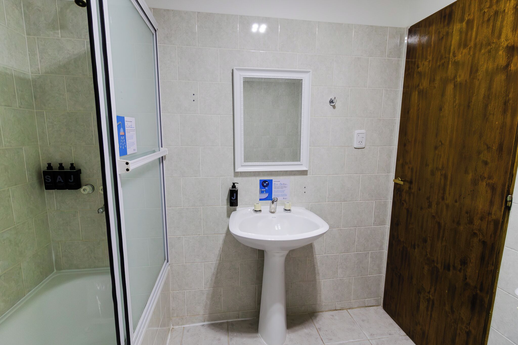 Un baño con ducha de vidrio, lavamanos blanco sobre pedestal y puerta de madera.