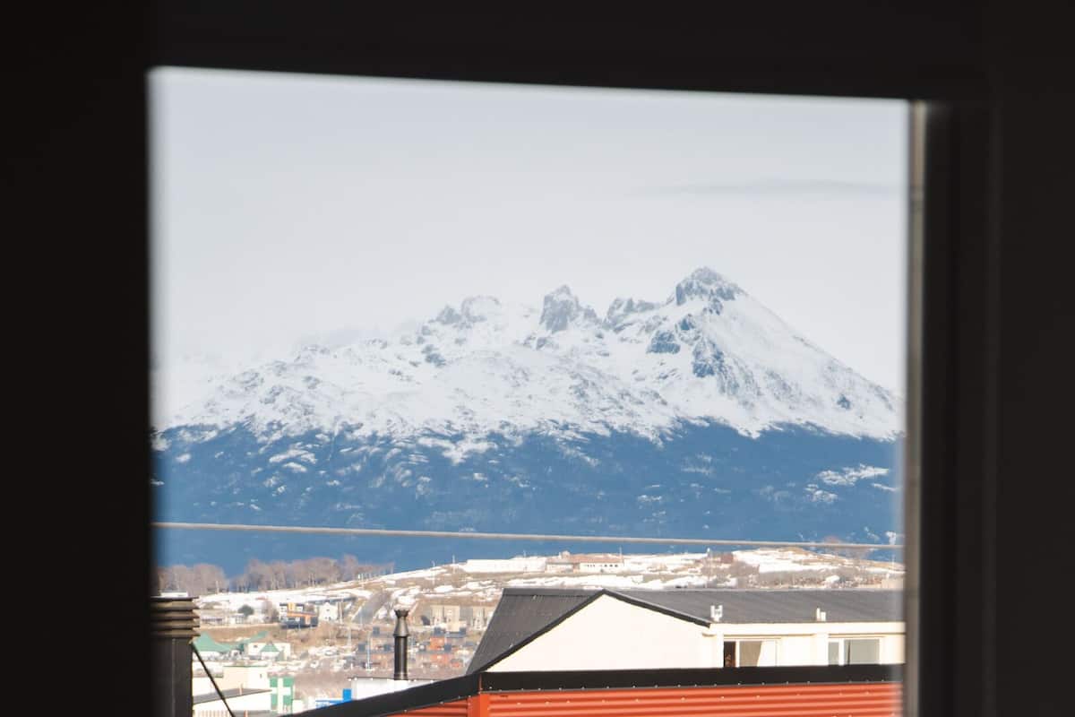 Una montaña con cima nevada, vista a través de una ventana con techo rojo y un edificio blanco al frente.