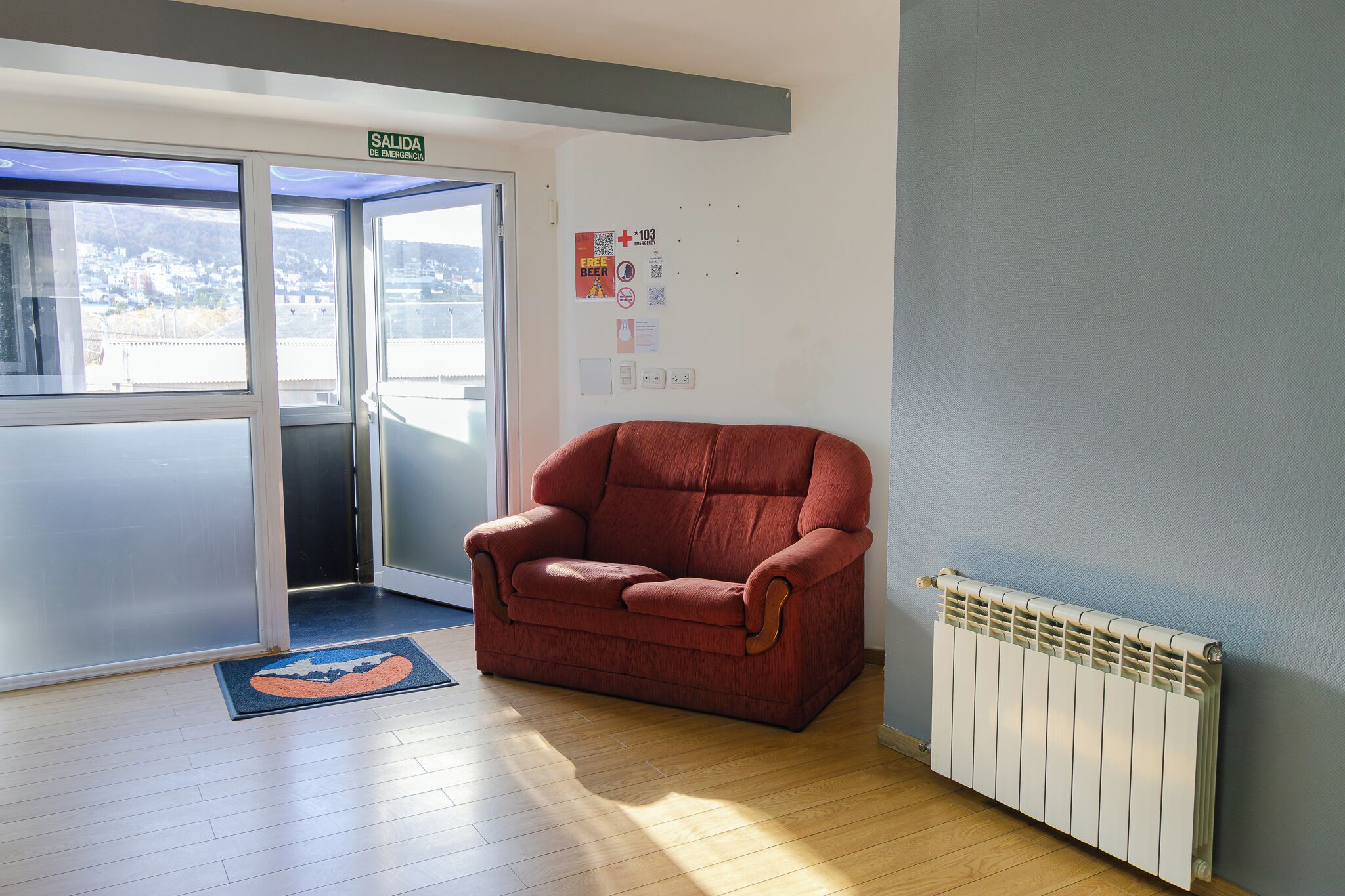 Une pièce avec un fauteuil rouge, un radiateur, une porte portant l’inscription « SORTIE » et une vue sur des immeubles, visible à travers la porte.