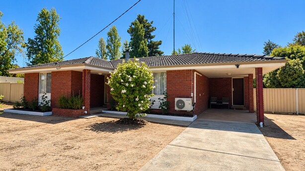 Casa de dos pisos con porche cubierto, fachada de ladrillo y un pequeño jardín con plantas florecientes.