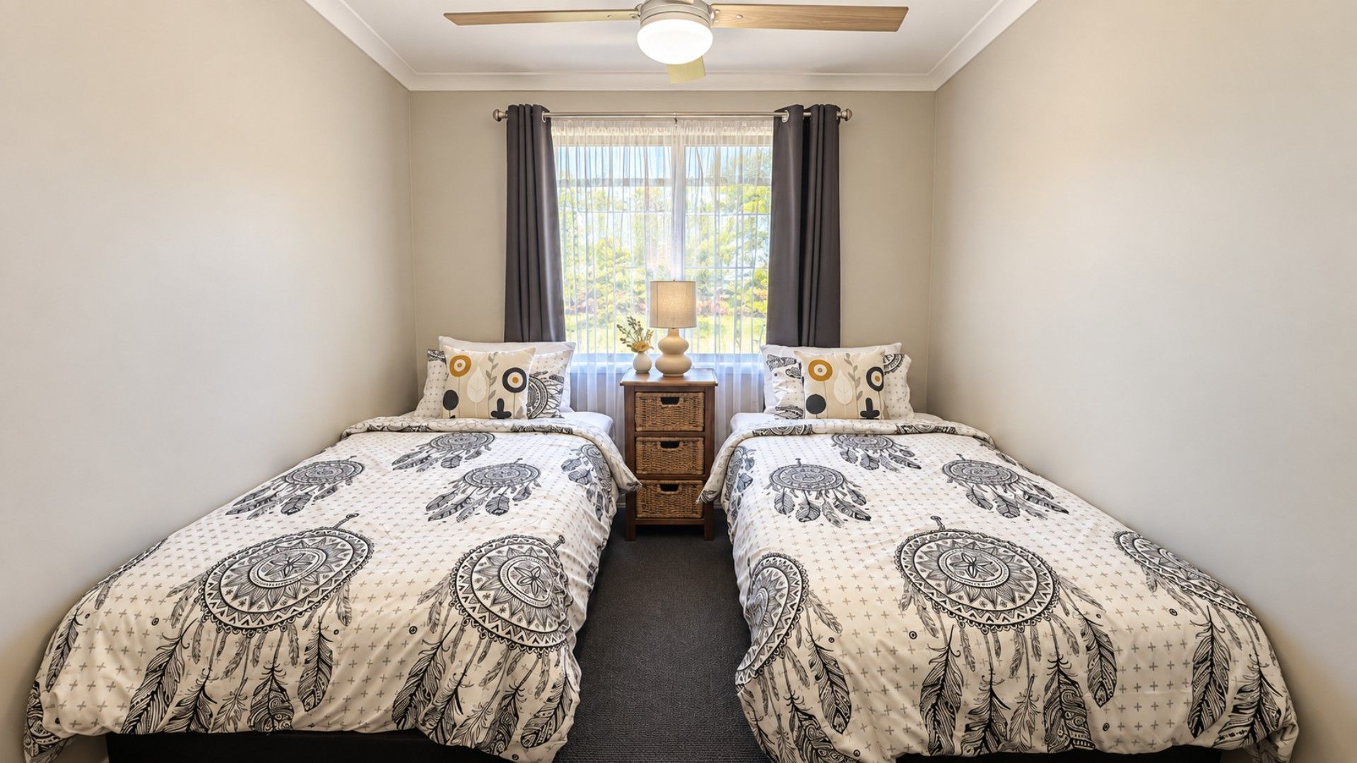 A bedroom with two beds, a ceiling fan, a window with curtains, and a bedside table with a lamp.