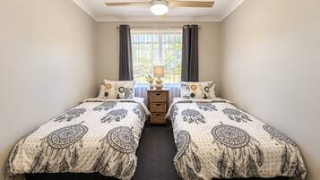 A bedroom with two beds, a ceiling fan, a window with curtains, and a bedside table with a lamp.