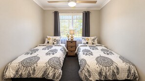 Un dormitorio con dos camas, un ventilador de techo, una ventana con cortinas y una mesita de noche con una lámpara.