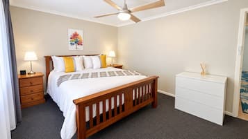 A bedroom with a wooden bed, two bedside tables with lamps, a ceiling fan, a white dresser, and a painting on the wall.