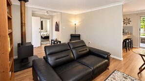 A modern living room with a black leather sofa, a wood-burning stove, a dining area with a table and chairs, and a view of the outdoors through a glass door.