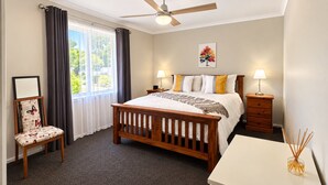 Un dormitorio con cama, mesitas de noche, un escritorio, una silla, ventilador de techo y ventana con cortinas.