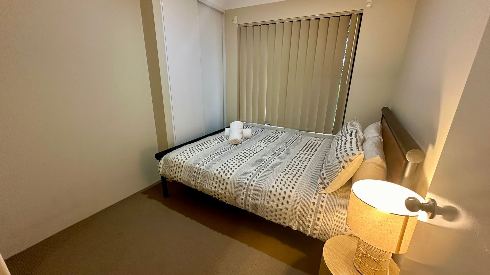 Een kleine, verzorgde slaapkamer met een enkel bed, een bedlampje en een raam met jaloezieën.