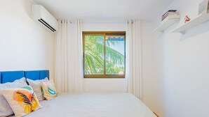 A bedroom with a bed, a window with curtains, a blue headboard, and an air conditioning unit. - IT81 Great 3/4 Apartment (1 Suite) - Itacimirim (Camaçari)