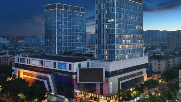 A modern high-rise building with a glass facade, surrounded by other buildings and a cityscape at dusk. - Homeinn · neo (Meishandong Railway Station Sansu Temple) (Meishan)