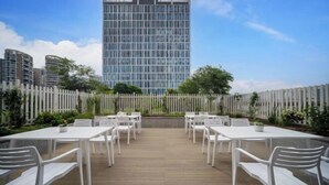 A rooftop terrace with white tables and chairs, a wooden deck, and a view of a tall building in the background. - Homeinn · neo (Meishandong Railway Station Sansu Temple) (Meishan)