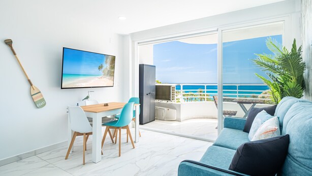 A modern living room with a large TV, a dining area, and a balcony with a view of the sea.