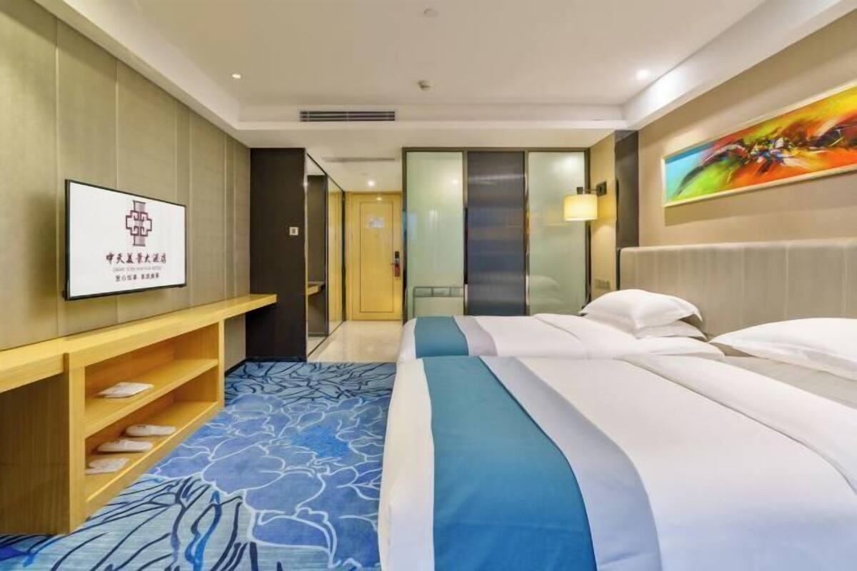 A hotel room with a bed, a TV on the wall, a wooden shelf with slippers, and a painting above the bed.