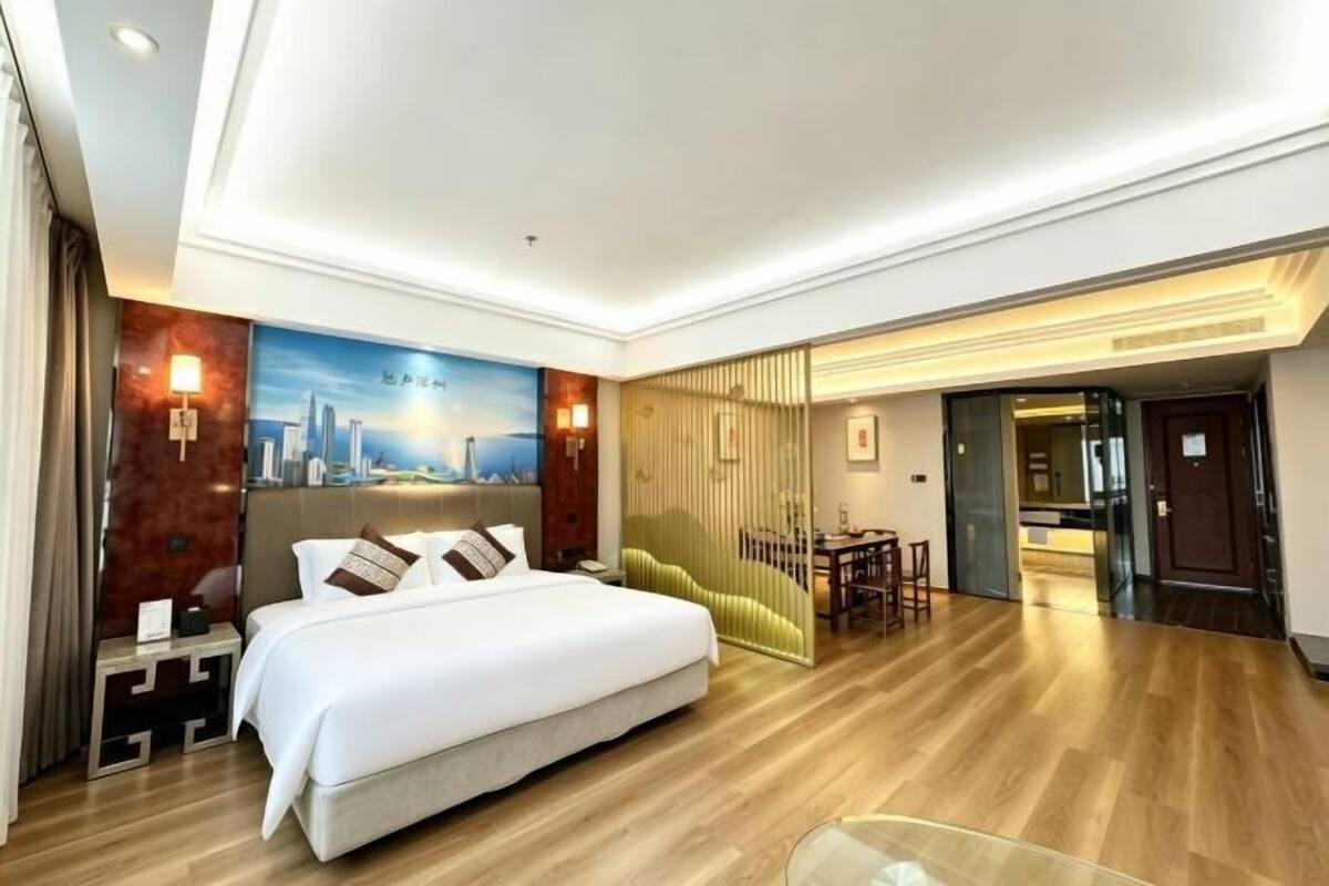 A hotel room with a large bed, a desk, a chair, a sofa, a dining area, and a view of the cityscape.