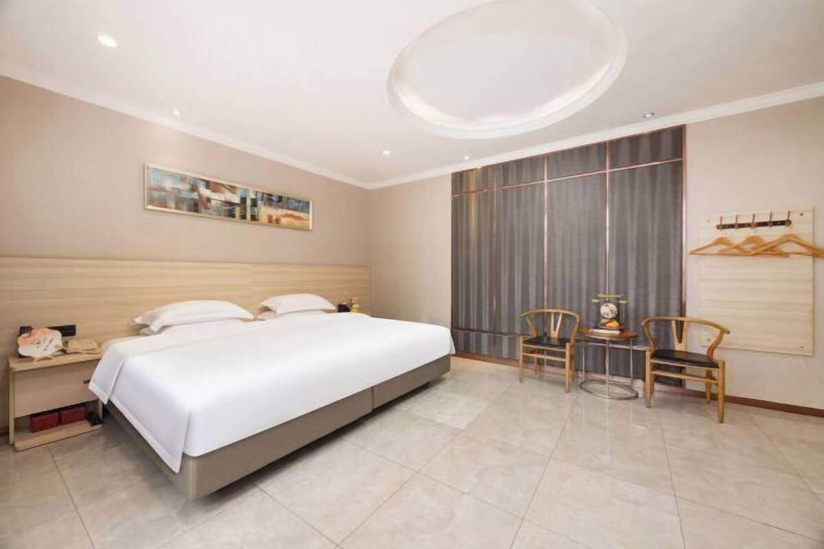 A hotel room with a bed, bedside table, chairs, a wall-mounted coat rack, and a large window with curtains.