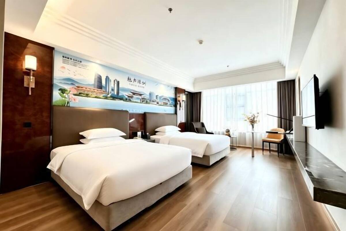 A hotel room with two beds, a flat-screen TV, a desk, and a large window.
