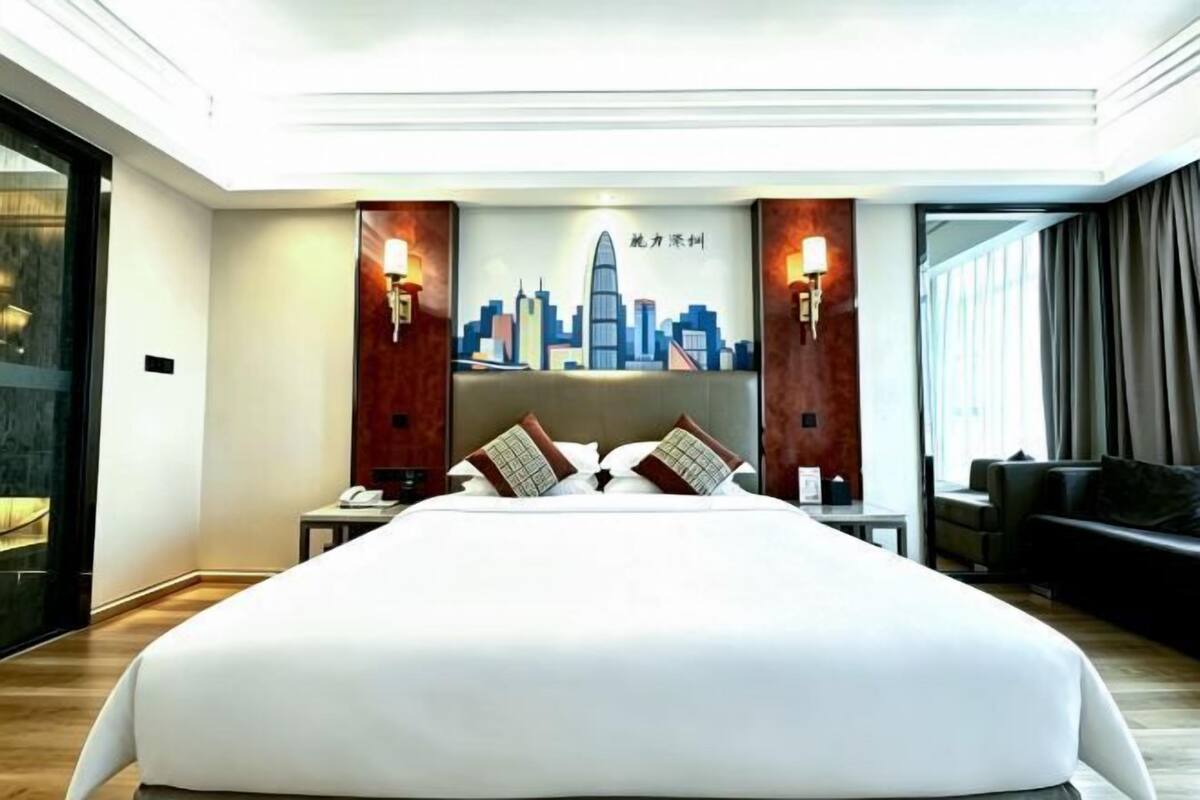A hotel room with a large bed, a cityscape artwork on the wall, a desk with a phone, and a sofa.