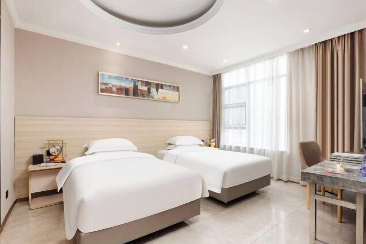 A hotel room with two beds, a desk, a chair, a painting on the wall, and a window with curtains.