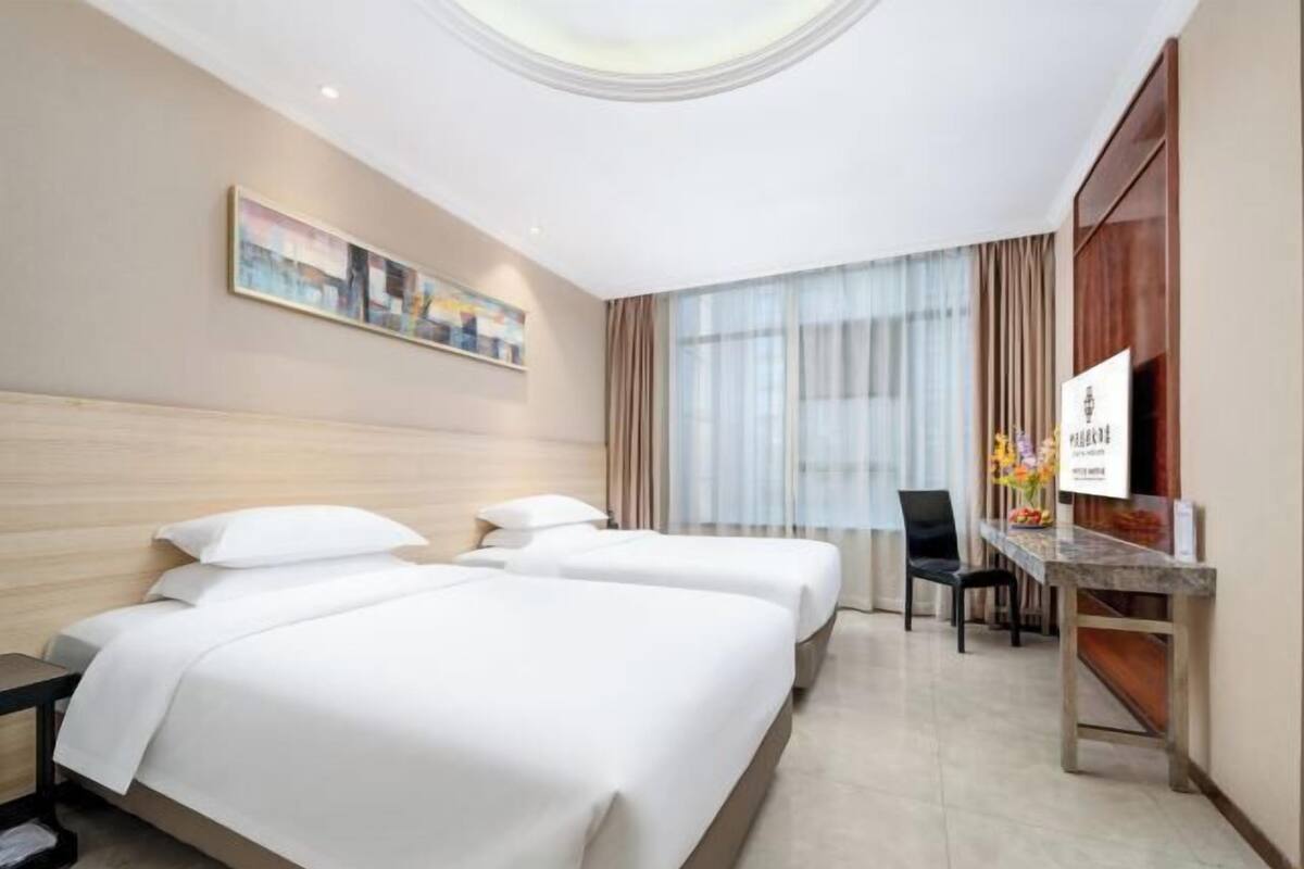 A hotel room with two beds, a desk, a chair, a painting on the wall, and a window with curtains.
