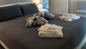 En pænt redt seng med mørkt sengetæppe, to puder med mønster og en stak hvide håndklæder.