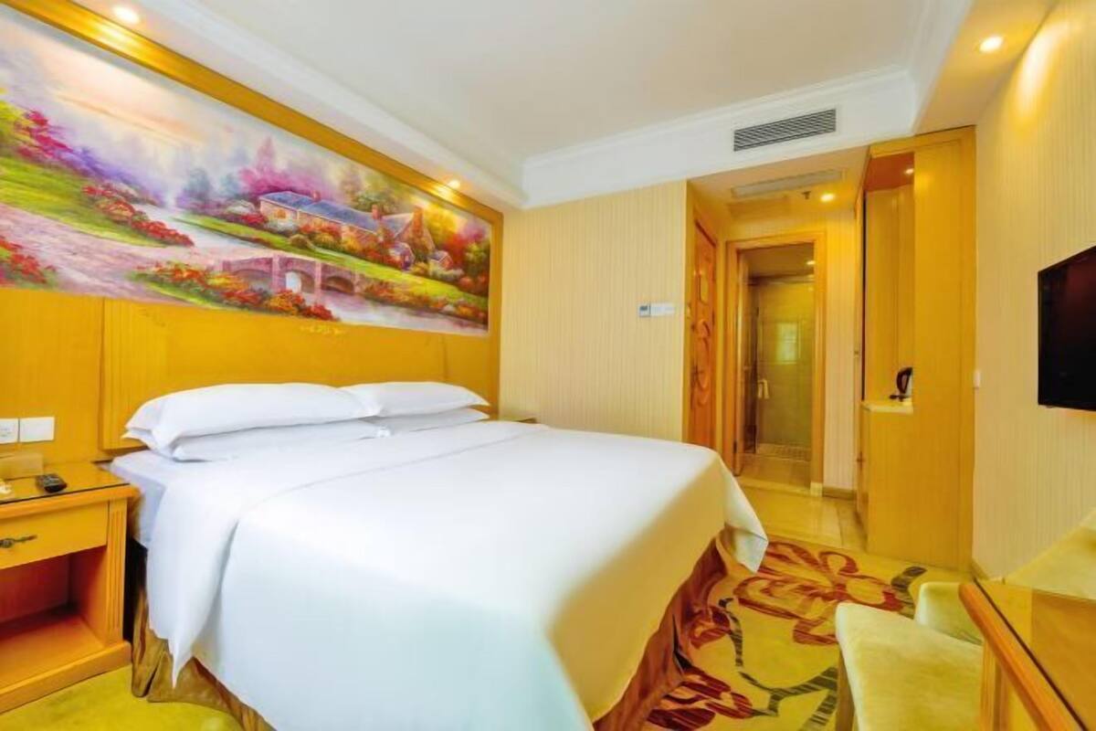 Ein Hotelzimmer mit einem großen Bett, einem Gemälde an der Wand, einem Fernseher und einem Holztisch.