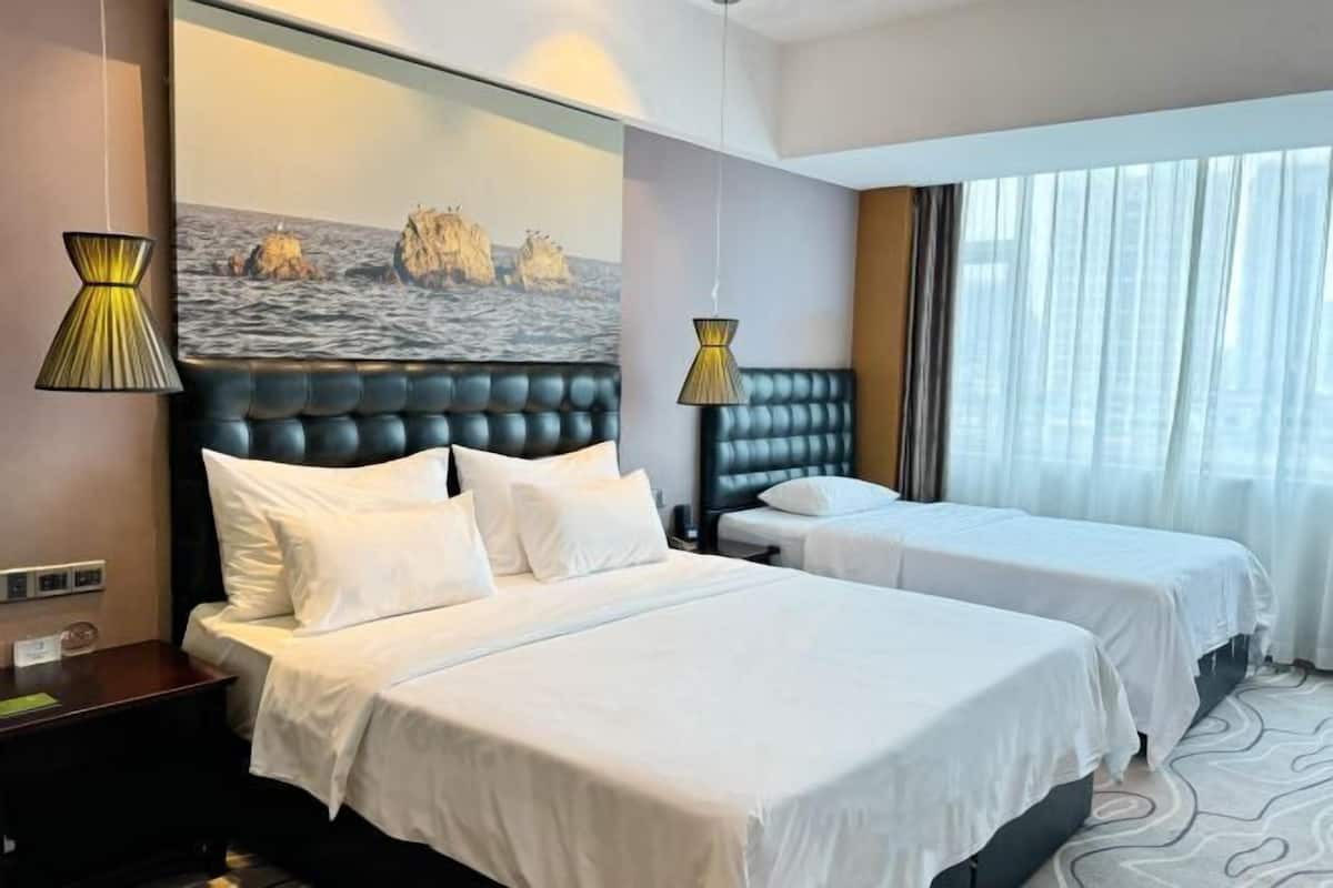 Ett hotellrum med två sängar, en stor sänggavel, en målning som föreställer ett kustlandskap och utsikt över staden genom stora fönster.
