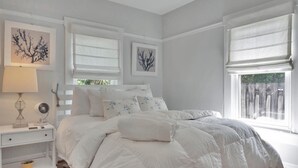 Ein Schlafzimmer mit einem großen Bett, weißen Nachttischen und zwei Fenstern mit Jalousien.