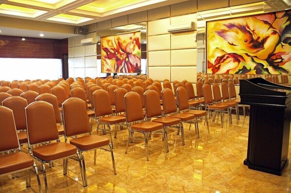 A conference room with rows of chairs, a podium, and large floral paintings on the walls. - Hotel Olympic (Jakarta)