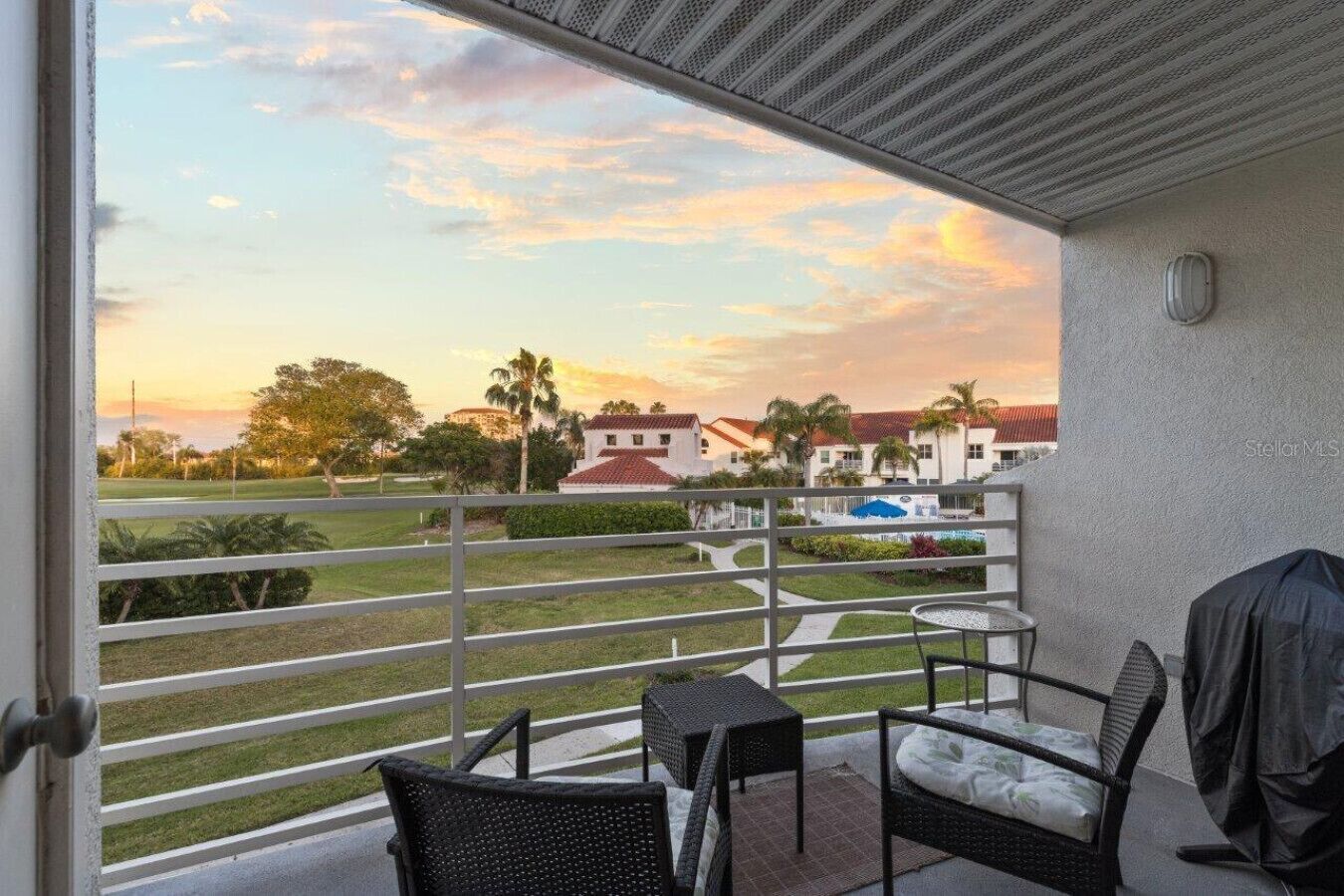 A balcony with wicker furniture, a grill, and a view of a golf course and buildings at sunset.