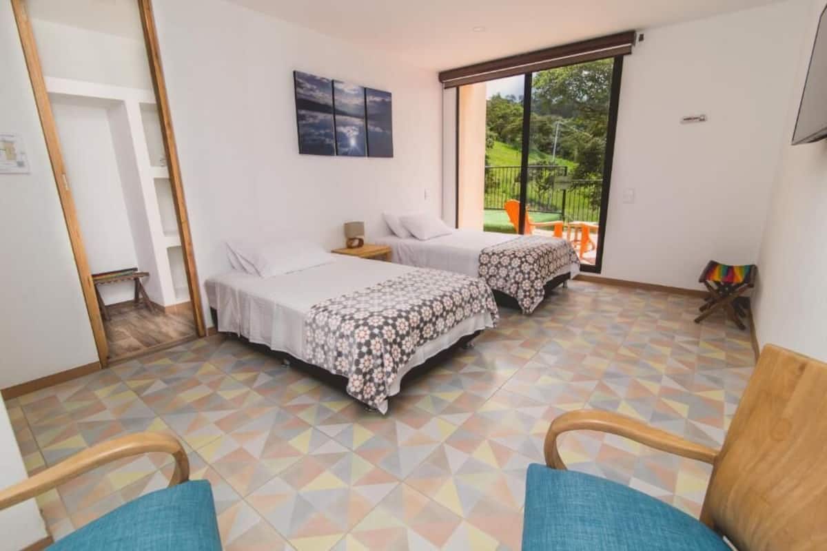 Quarto com duas camas, uma mesinha, uma cadeira e varanda com vista para o verde.