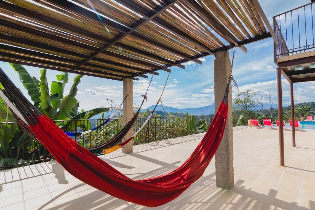 Een hangmat onder een houten pergola met uitzicht op bergen en een zwembadgedeelte met ligbedden.