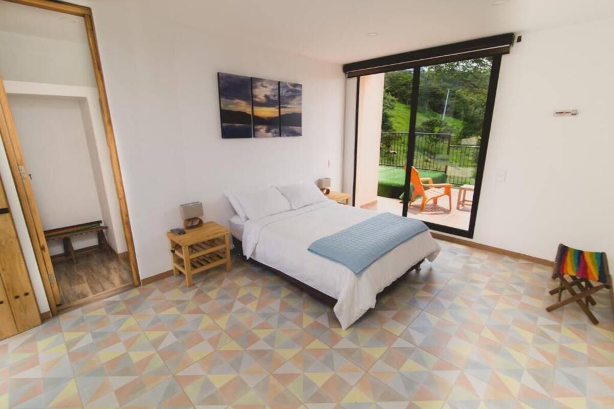 Quarto com cama, mesa de cabeceira, janela com vista para vegetação e uma cadeira colorida.