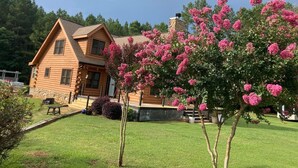 A log cabin with a stone foundation, a well-maintained lawn, and a flowering tree in the foreground.