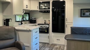 A compact kitchen with white cabinets, a microwave, a stove, and a refrigerator. There is a seating area with a sofa and a chair.