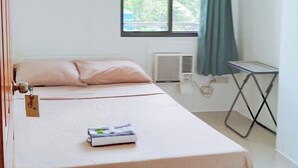 Una cama bien hecha con una etiqueta, una ventana con cortinas, un radiador y una mesita.