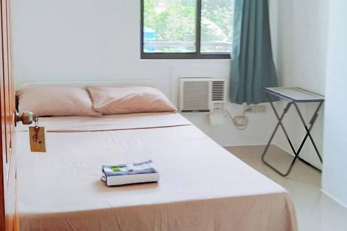 Een keurig opgemaakt bed met een label, een raam met gordijnen, een radiator en een klein tafeltje.