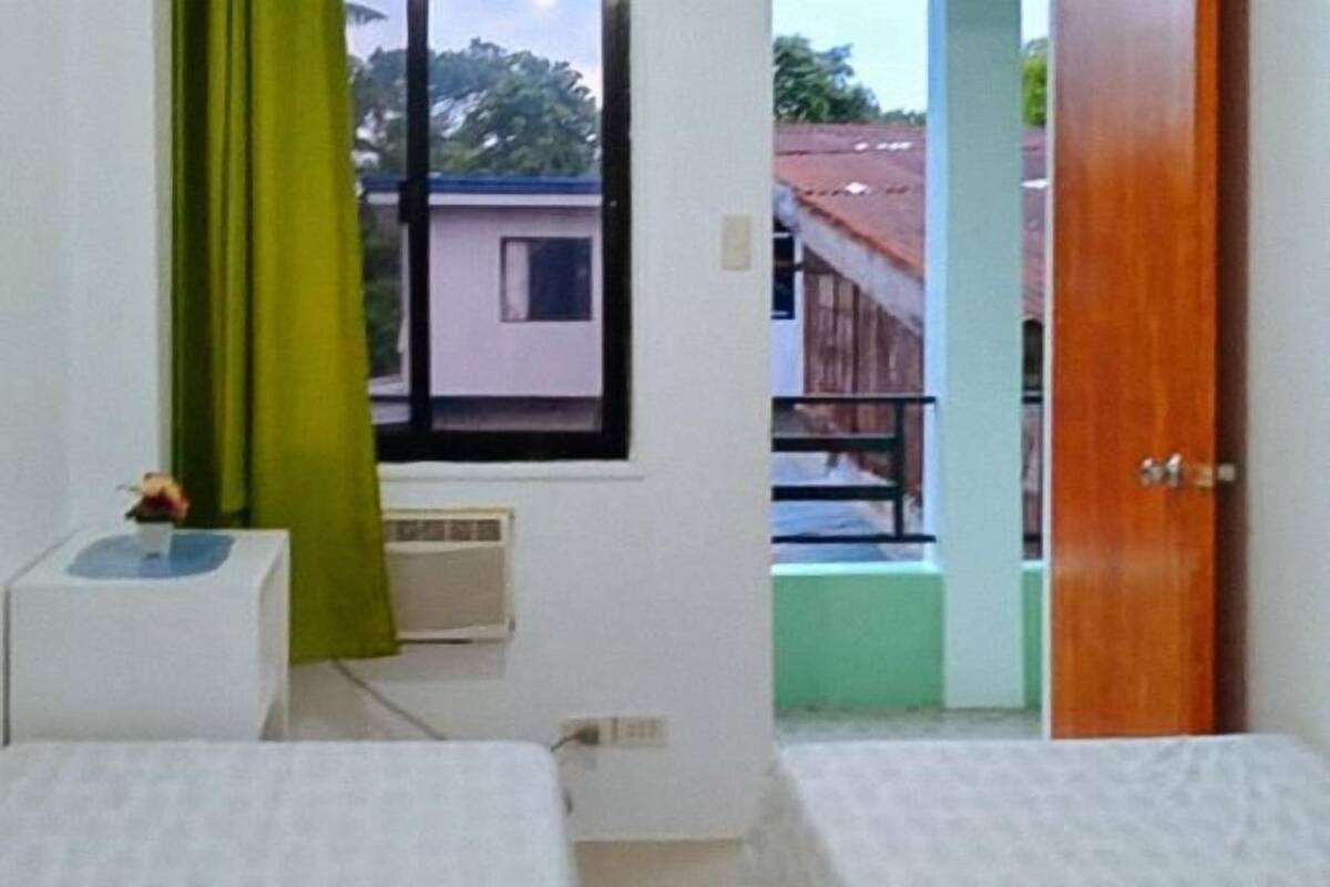 Een kleine kamer met een bed, een raam met uitzicht op gebouwen, een deur en een tafeltje met een bloem.