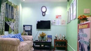 Una sala de estar con un sofá, un televisor montado en la pared, una mesita con un jarrón de flores y un cartel que dice 'Welcome'.