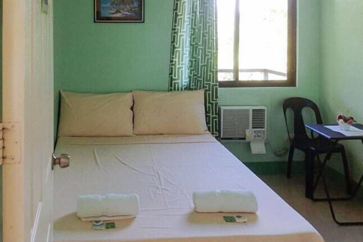 Een enkel bed met twee kussens, een nachtkastje met een stoel, een raam met gordijnen met een patroon, en een luchtverkoeler aan de muur.